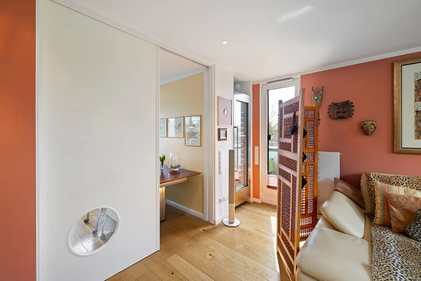 Interior view of a living room with a white sofa, wooden screen, and a sliding door with an oval window. The walls are painted in peach and white.