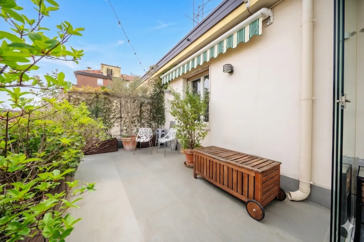 A rooftop patio with green plants, two white chairs, and a wooden bench with wheels. The sky is blue.