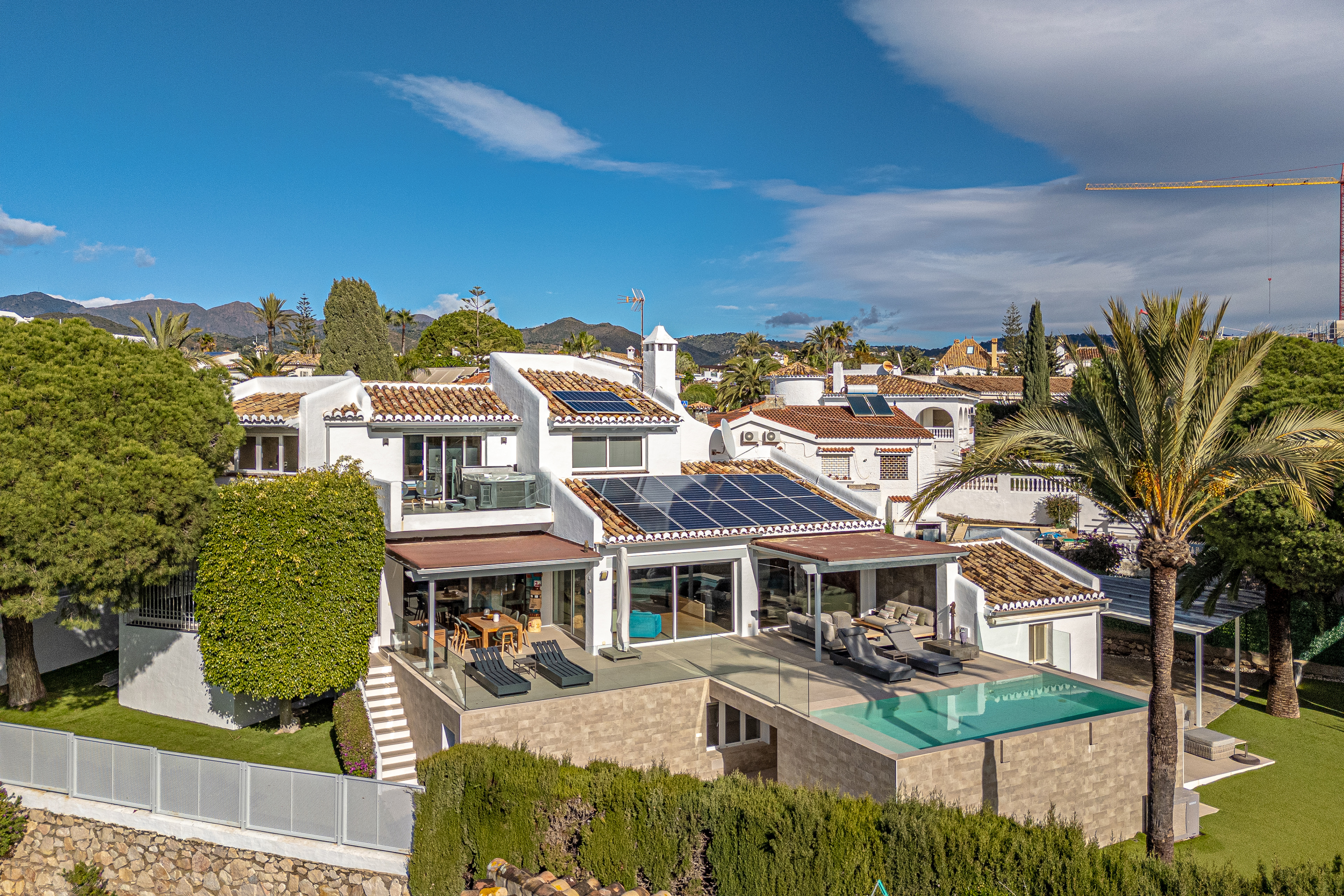 Aerial view of a white villa with a pool, solar panels, and a palm tree against a blue sky.