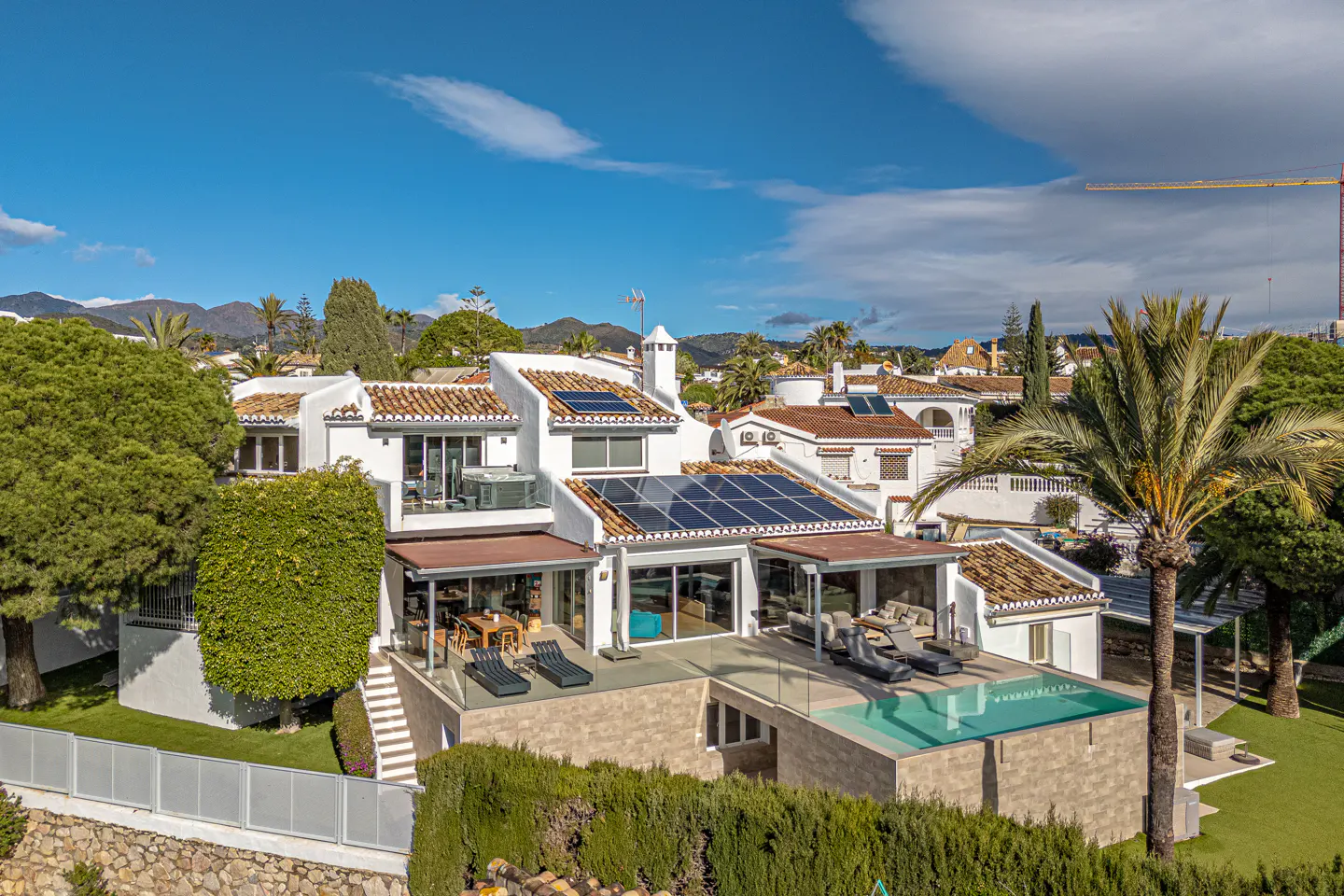 Aerial view of a white villa with a pool, solar panels, and a palm tree against a blue sky.