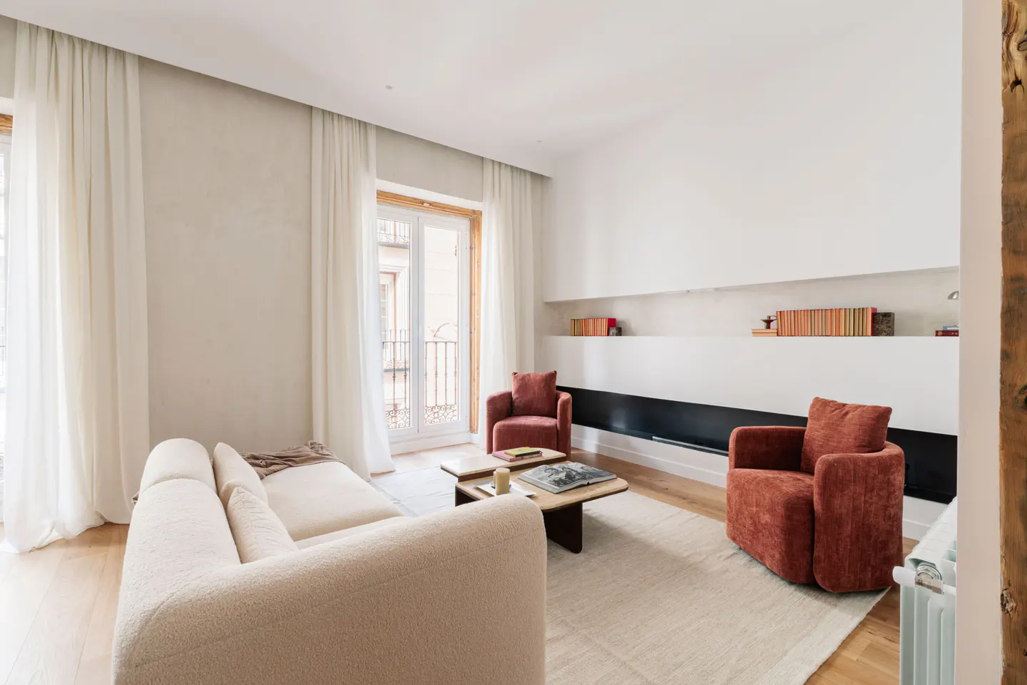 Bright living room with a cream sofa, two rust-colored armchairs, and a light rug. White walls with built-in shelves and a black accent. Balcony doors with white curtains.
