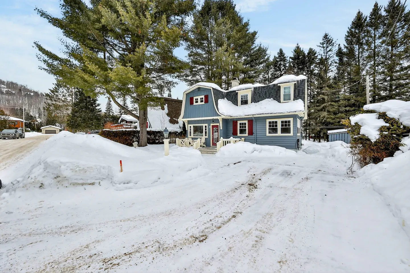 A blue house with red shutters is surrounded by snow in a winter landscape. Tall trees are in the background.