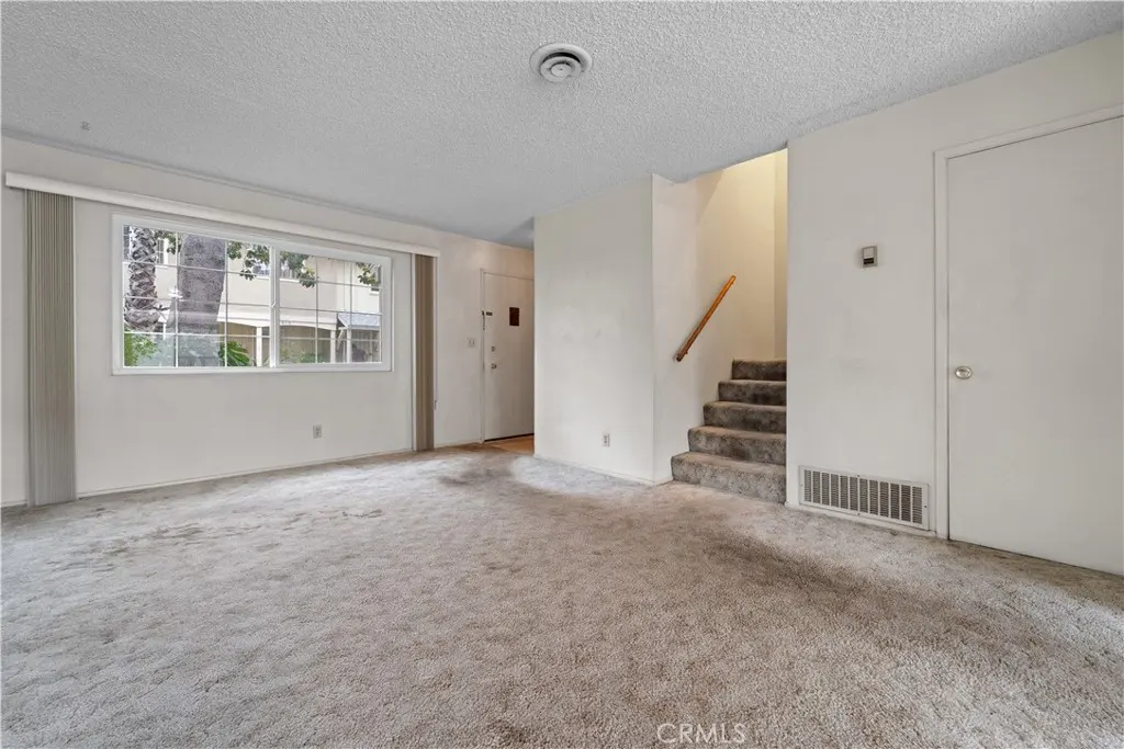 A bright, empty living room with white walls, gray carpet, a window, and stairs.