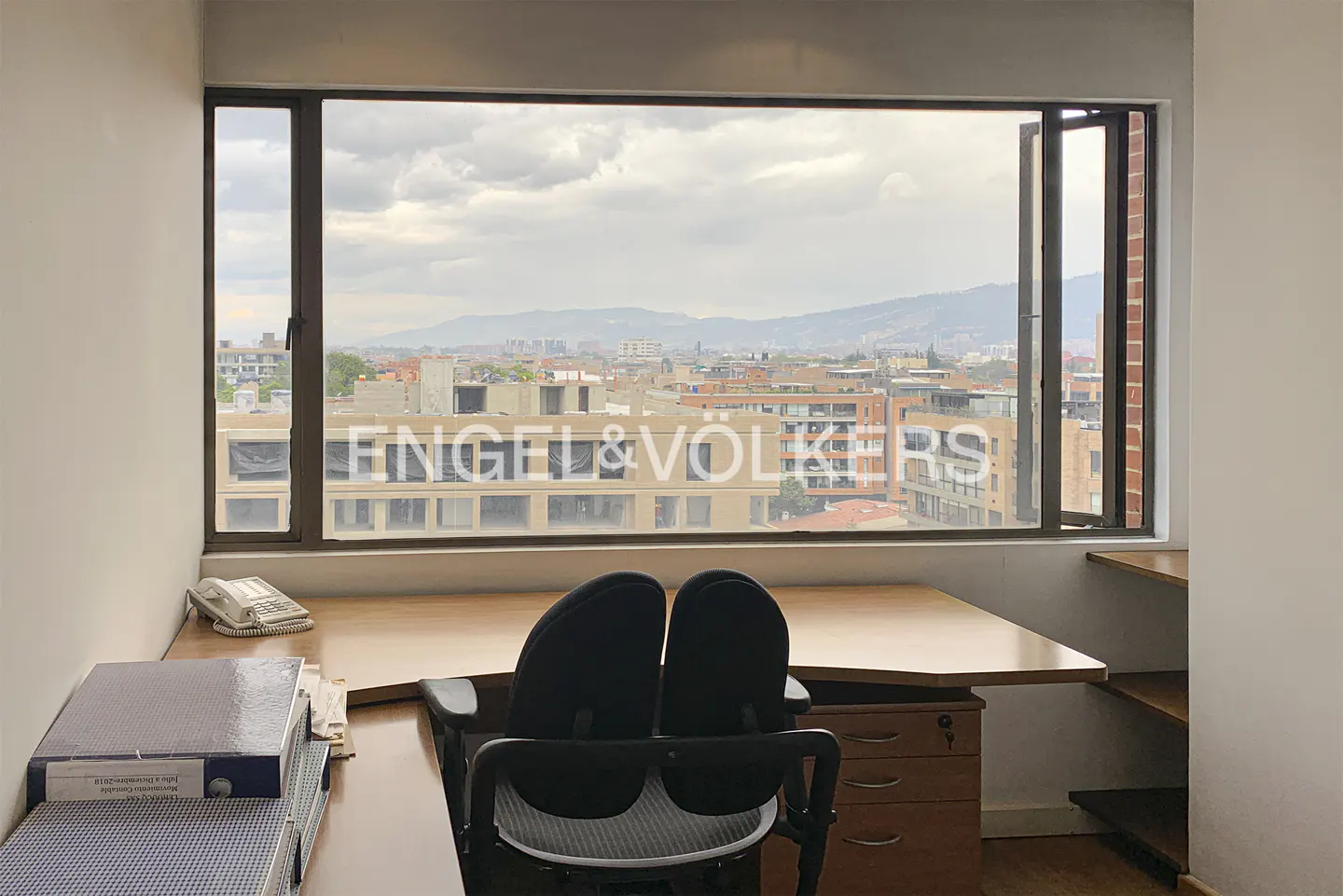 Office space with a desk, chair, and a large window overlooking a cityscape with mountains in the distance. Engel & Völkers logo visible.