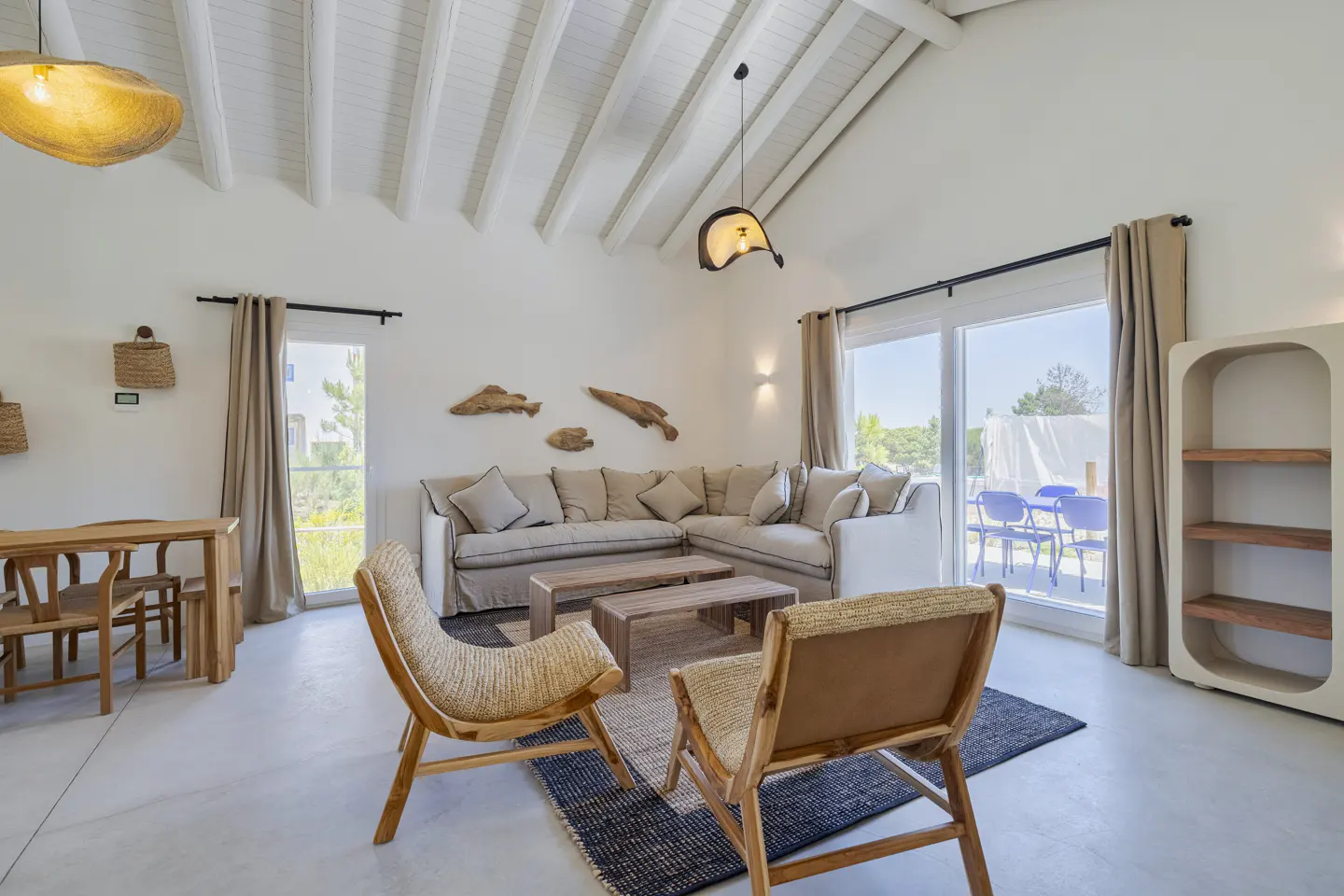 Bright living room with white vaulted ceilings, beige sectional sofa, two wooden chairs, and a view of a patio.