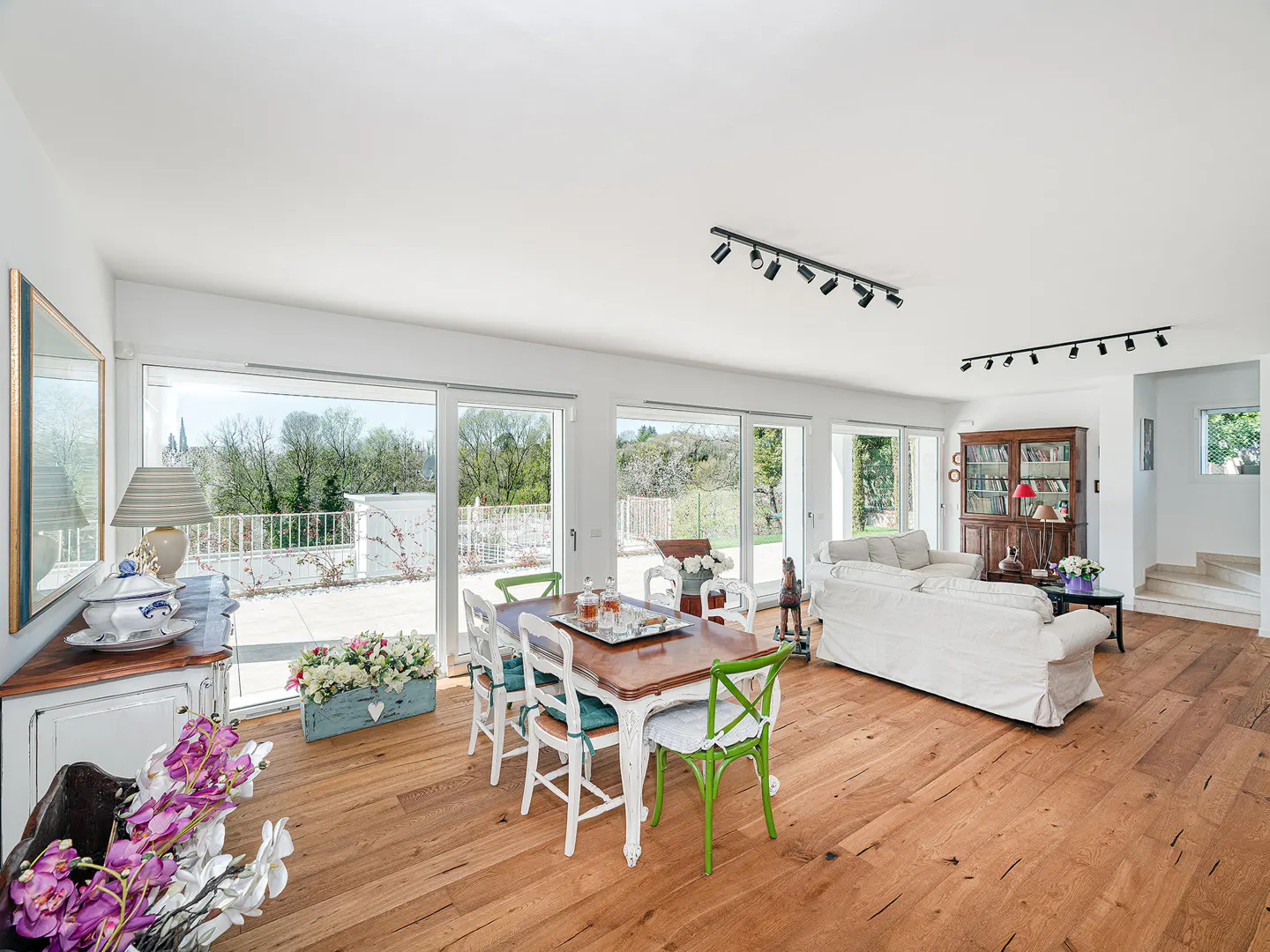 Bright, open-plan living and dining area with wood floors, white walls, and large windows overlooking a green landscape. Dining table with colorful chairs.