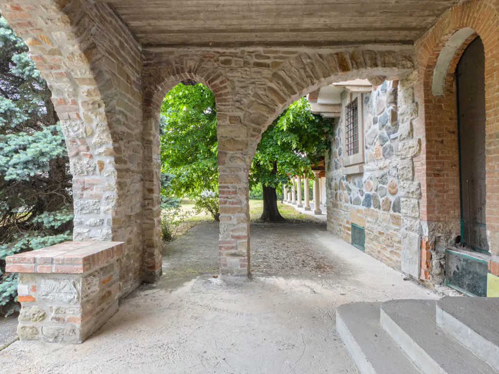 Covered stone patio with arched columns, concrete steps, and a view of a lush green tree in the garden.