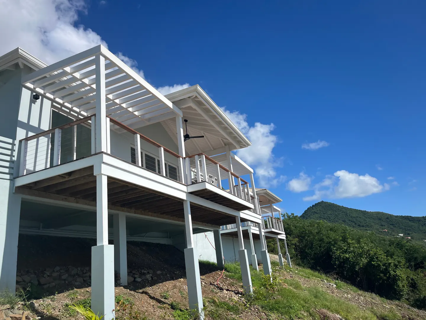Exterior view of a light blue house with white decks and a pergola, set against a blue sky and green hillside.