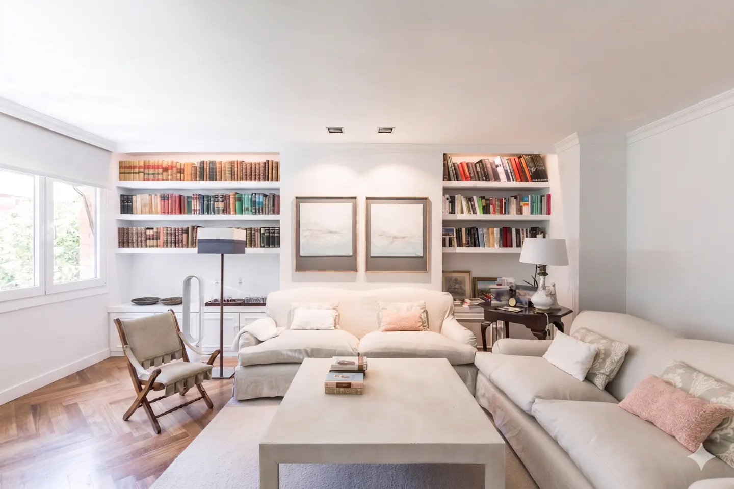Bright living room with two cream sofas, a coffee table, a wooden chair, and built-in bookshelves filled with books.