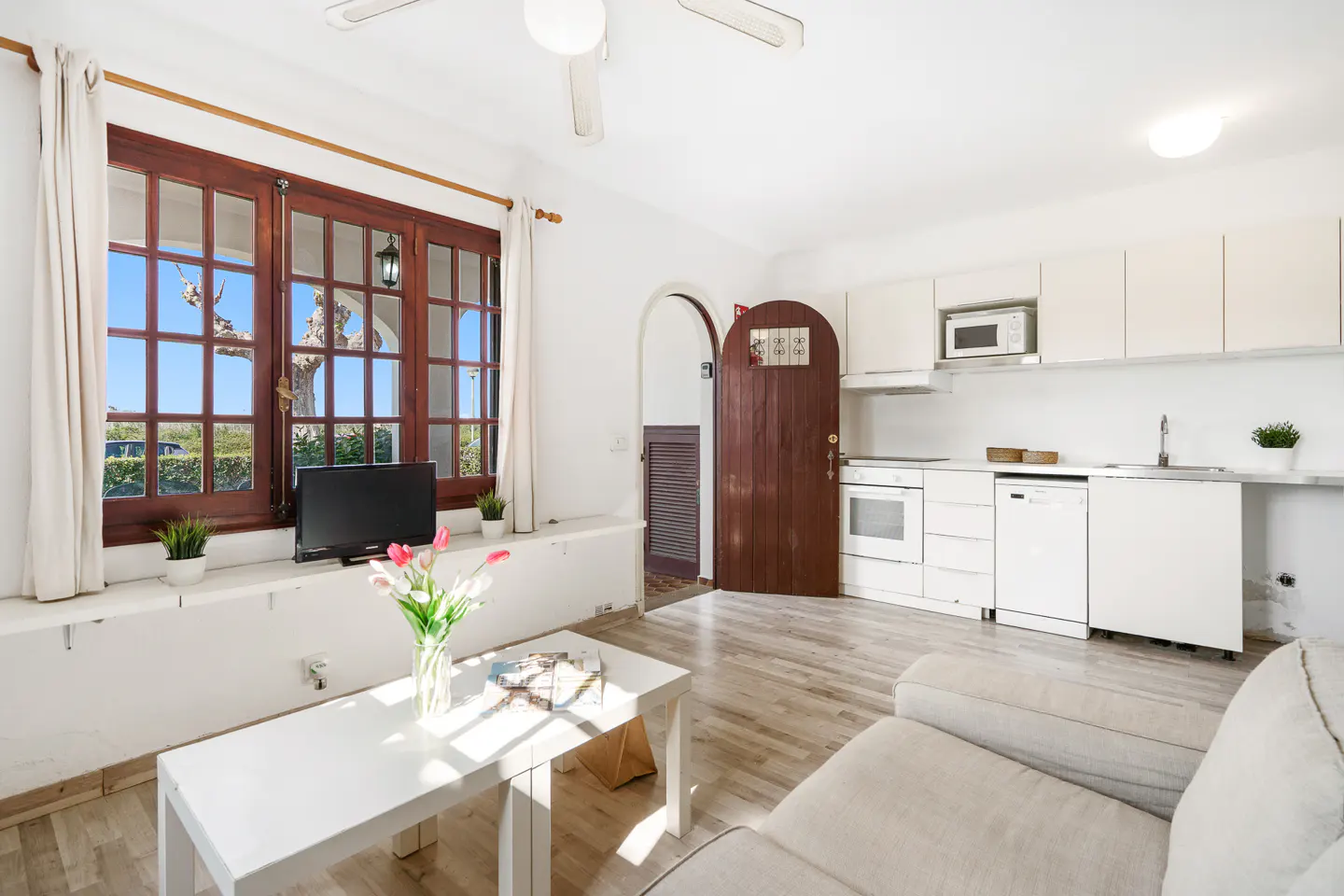 Bright, open-concept living space with white walls, wood floors, and a white sofa. A white kitchen is visible, and a window with brown trim lets in natural light.