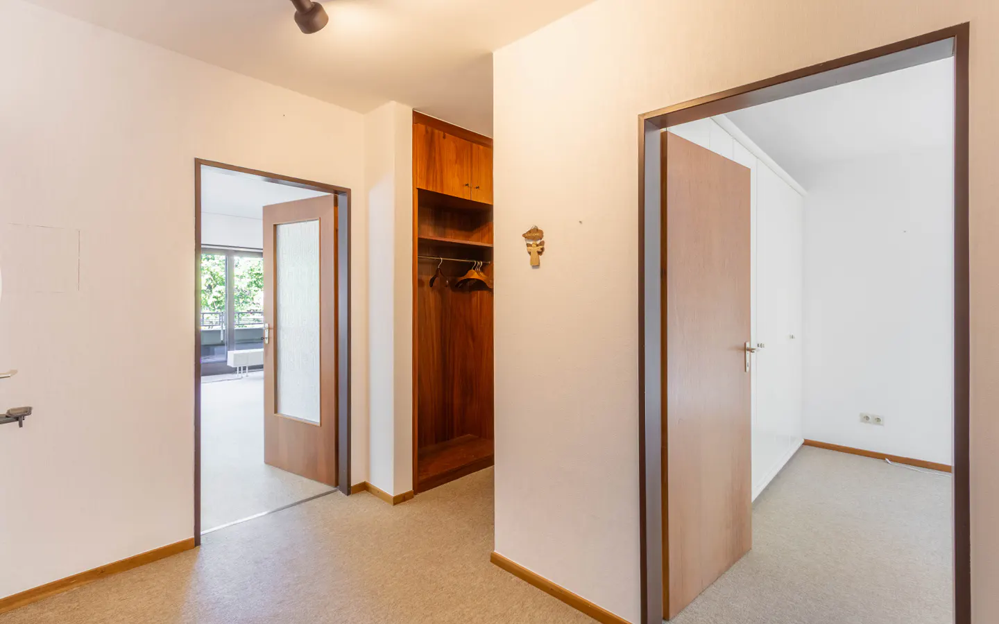 Hallway with light carpet, a wooden open closet, and two doorways leading to bright rooms with wooden doors.
