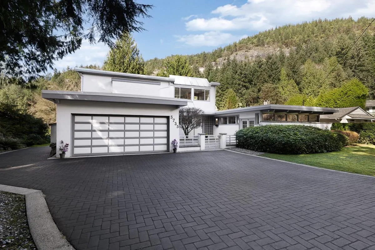 Modern white house with a gray garage door and brick driveway, set against a backdrop of lush green trees and a mountain.