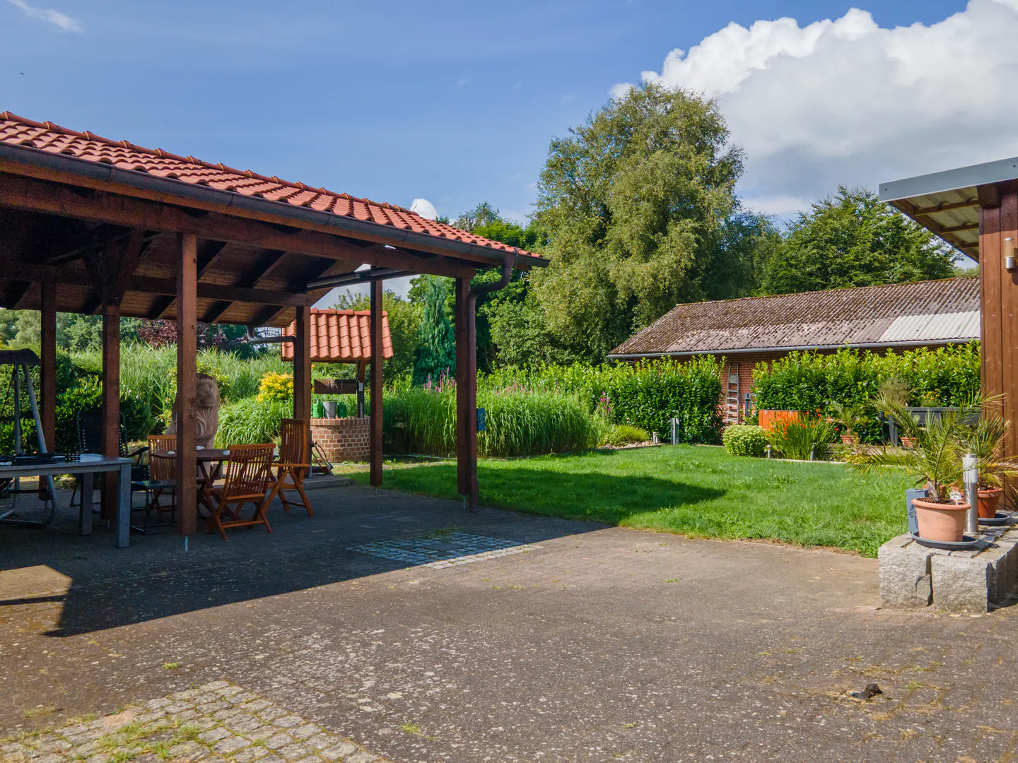 Outdoor patio with wooden pergola, table, and chairs. A brick well and green lawn are in the background.