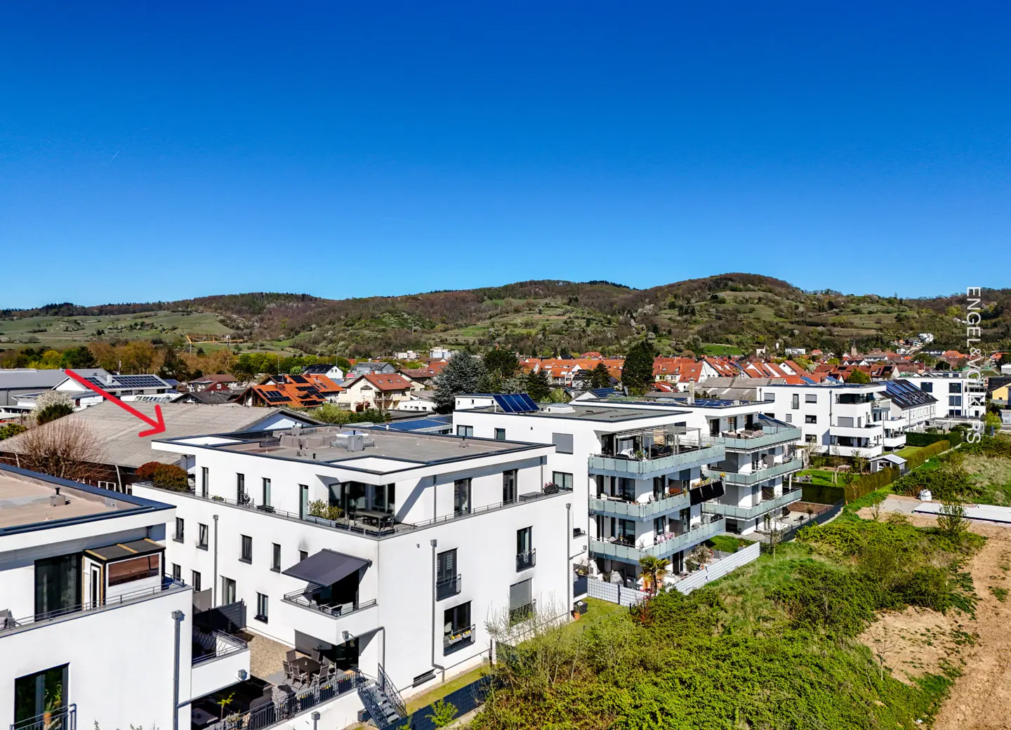 Aerial view of modern white apartment buildings with balconies, green hills, and a clear blue sky.