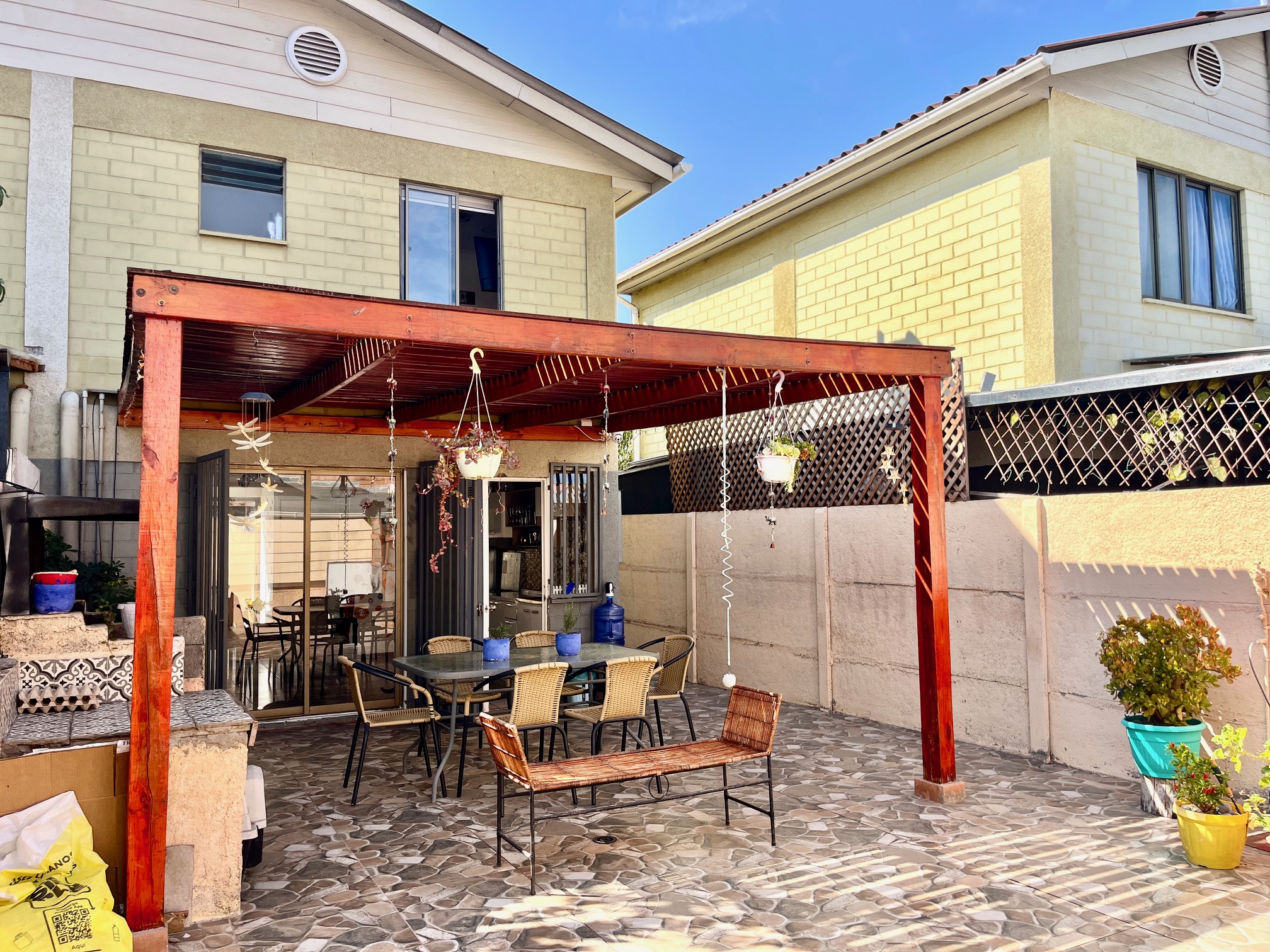 Outdoor patio with a red pergola, table, chairs, and bench on a stone tile floor. Potted plants and hanging baskets add greenery.