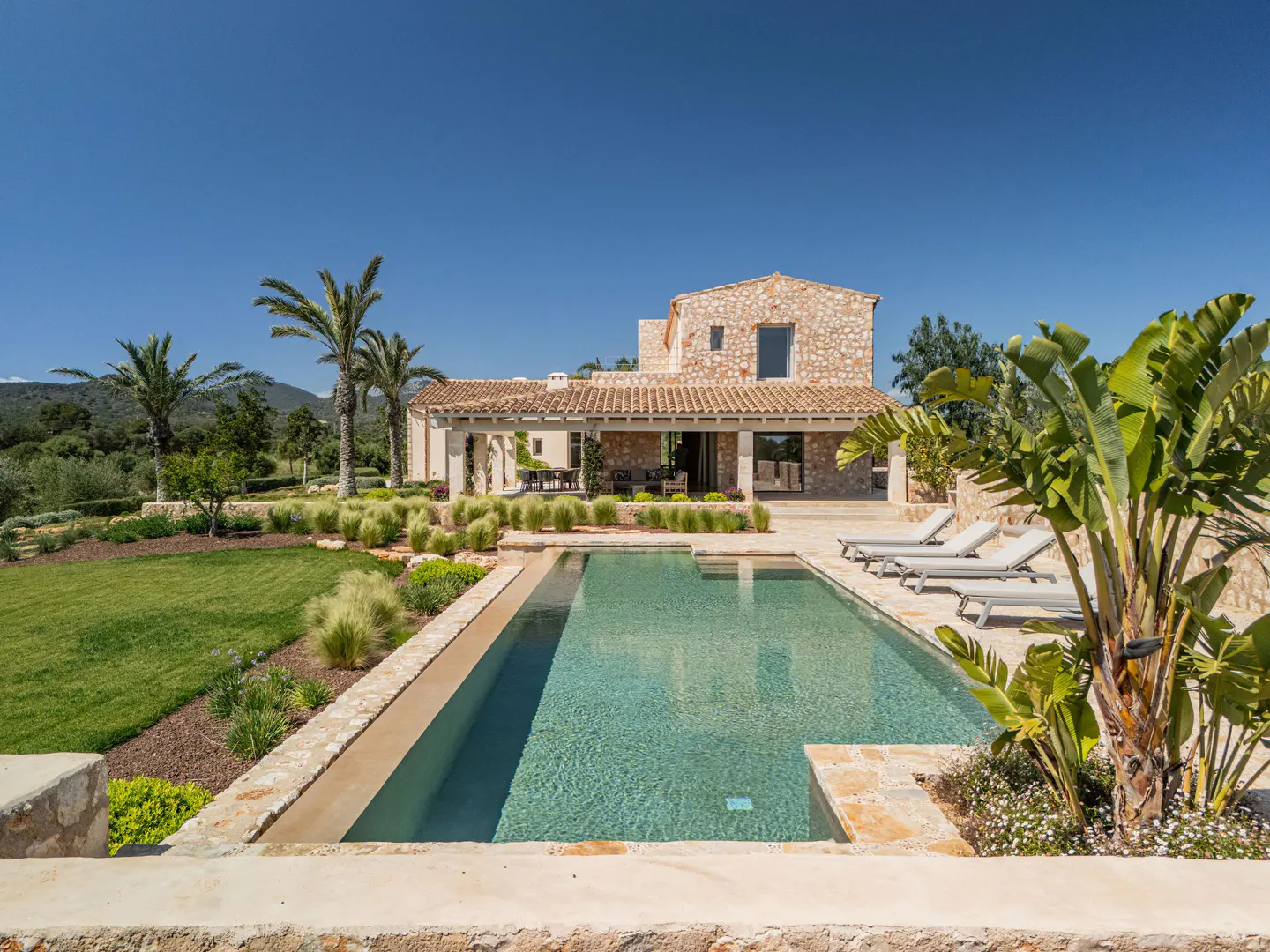 A stone villa with a swimming pool, lounge chairs, palm trees, and a green lawn under a clear blue sky.