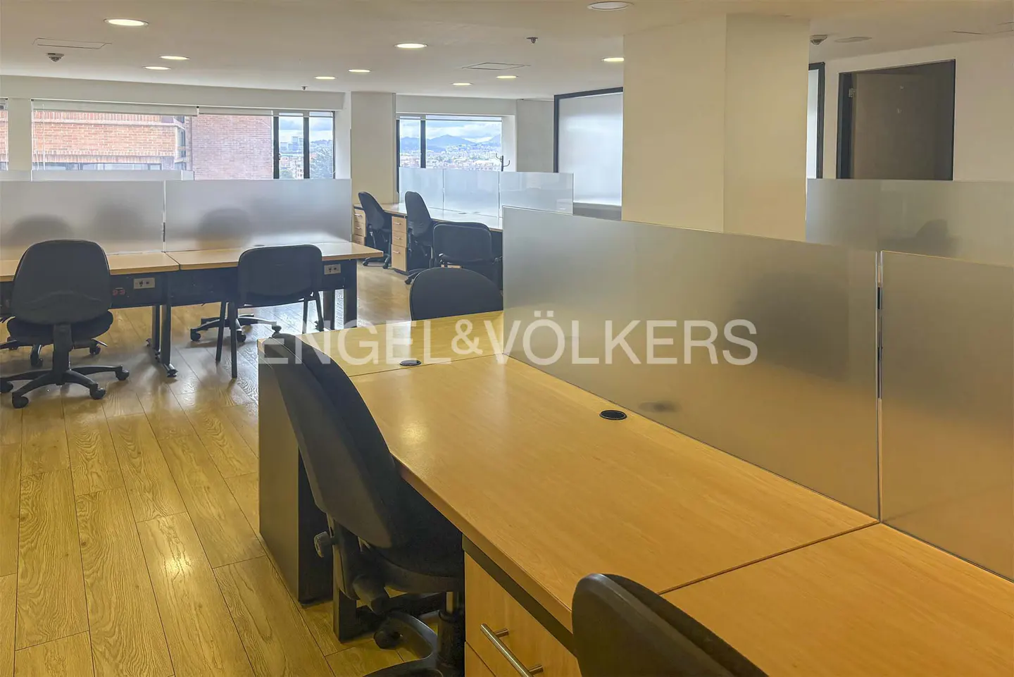 Modern office space with light wood desks, black ergonomic chairs, and frosted glass partitions on a wooden floor.