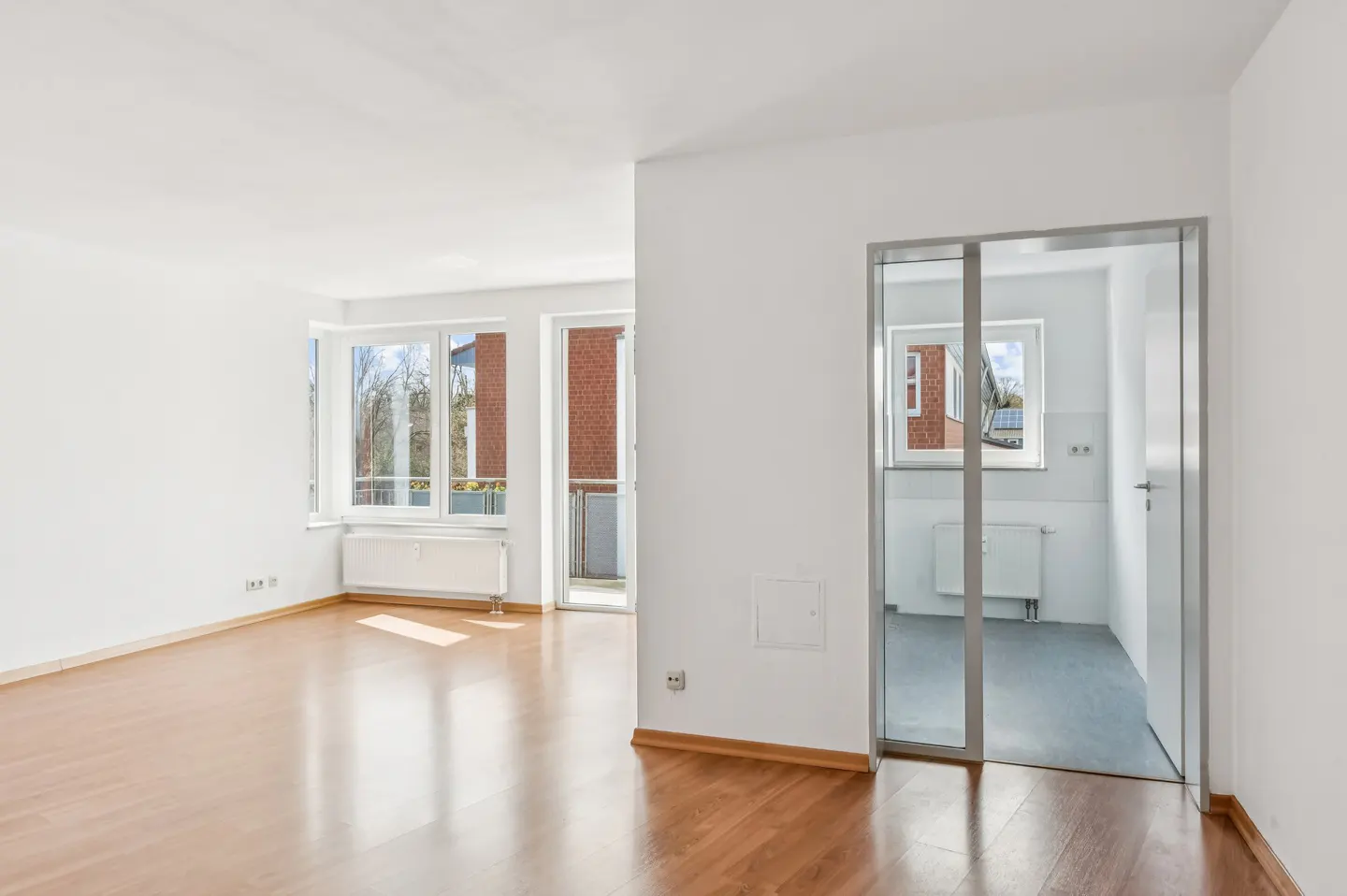 Bright, empty room with wood floors, white walls, and large windows. A glass door leads to another room. Outside, a brick building is visible.