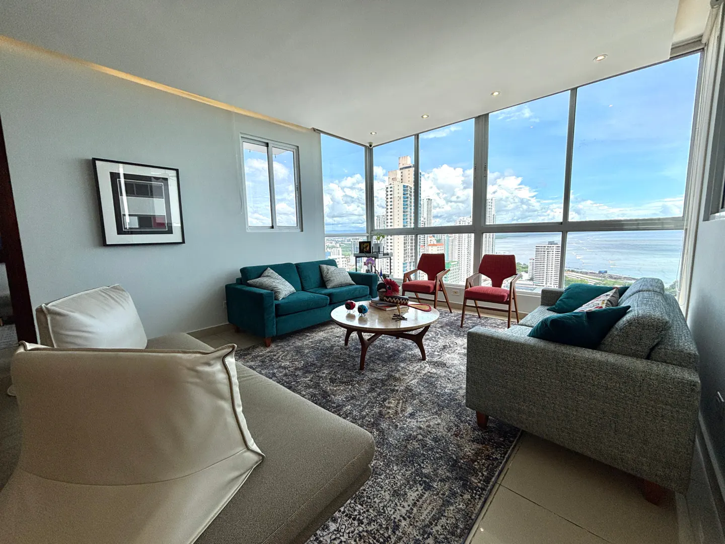 Bright living room with teal sofa, gray sofa, and two red chairs. Large windows offer a city and ocean view.