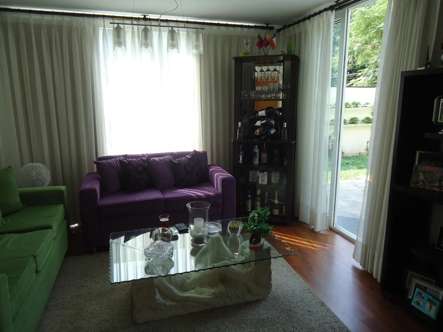 Living room with a green sofa, a purple sofa, a glass coffee table, and a dark wood cabinet with glass doors. White curtains cover the windows.
