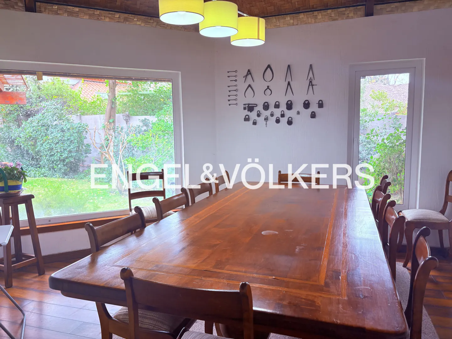 A dining room with a long wooden table and chairs, a window overlooking a green garden, and a wall with decorative metal objects.