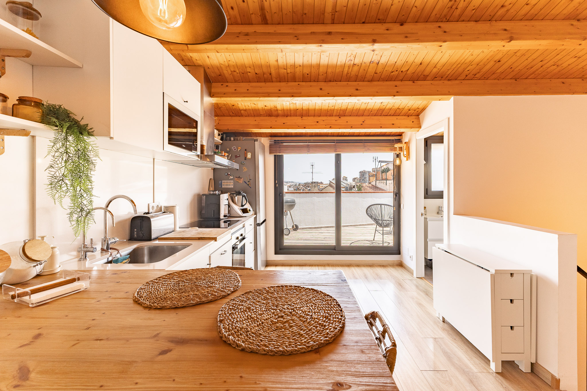 Modern kitchen with wooden ceiling beams, white cabinets, a wooden dining table, and a sliding door leading to a balcony.