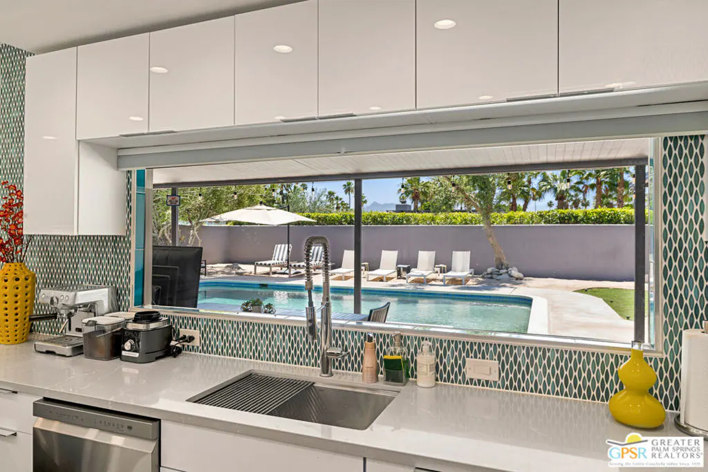 A kitchen with white cabinets and a pass-through window overlooking a pool with lounge chairs.