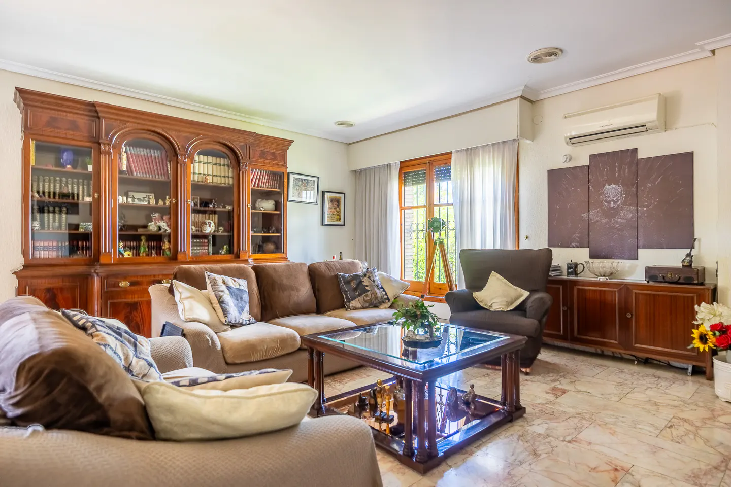 Living room with brown sofas, armchair, glass coffee table, and a large wooden bookcase. Marble floor and natural light.