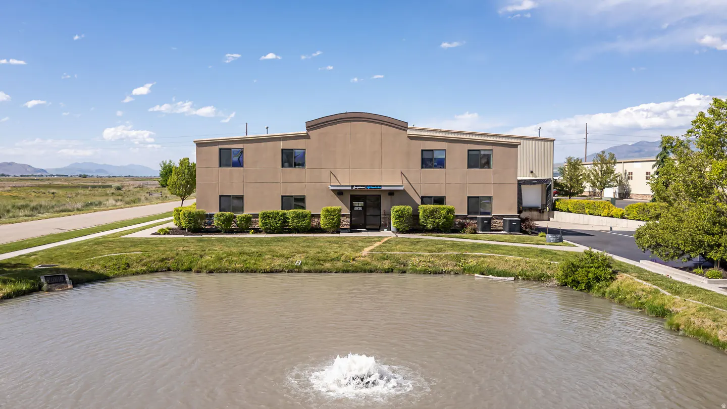 Tan commercial building with a pond and fountain in front, green grass, and blue sky.