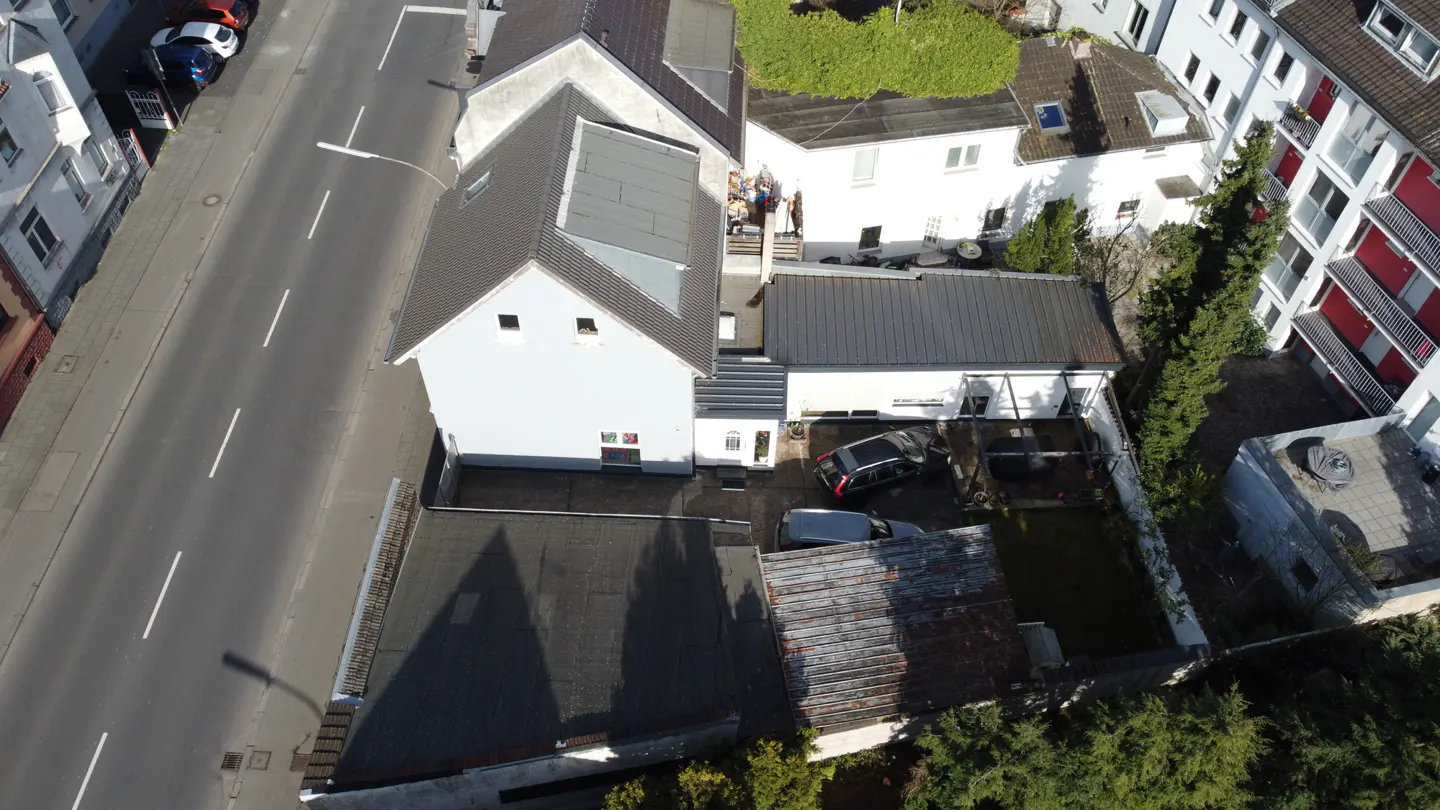 Aerial view of a white house with a gray roof, a black roof, and a green lawn, with cars parked in the driveway.