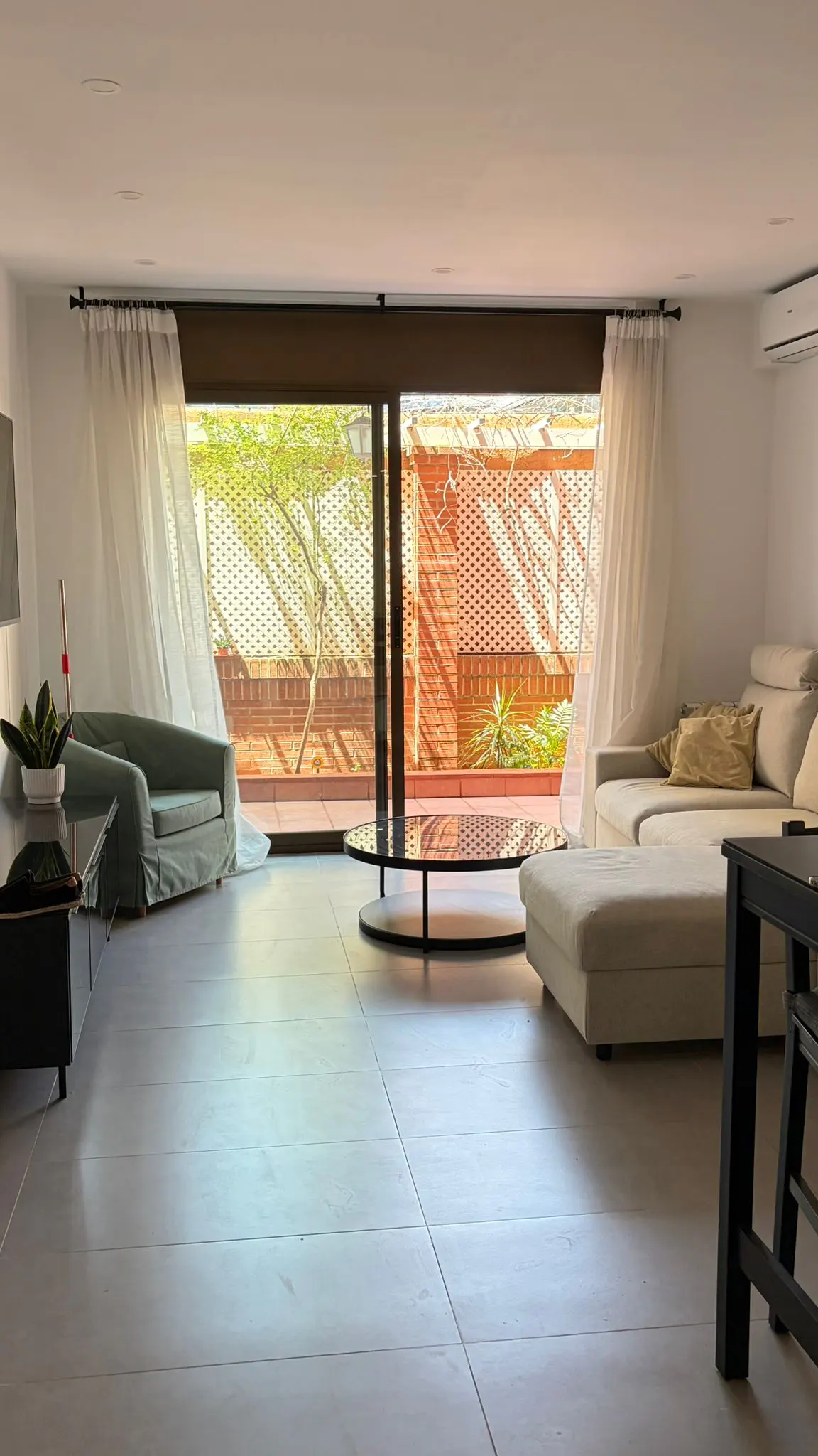 Bright living room with a green armchair, a cream sofa, and a round coffee table. Large sliding glass doors lead to a patio with a brick wall.