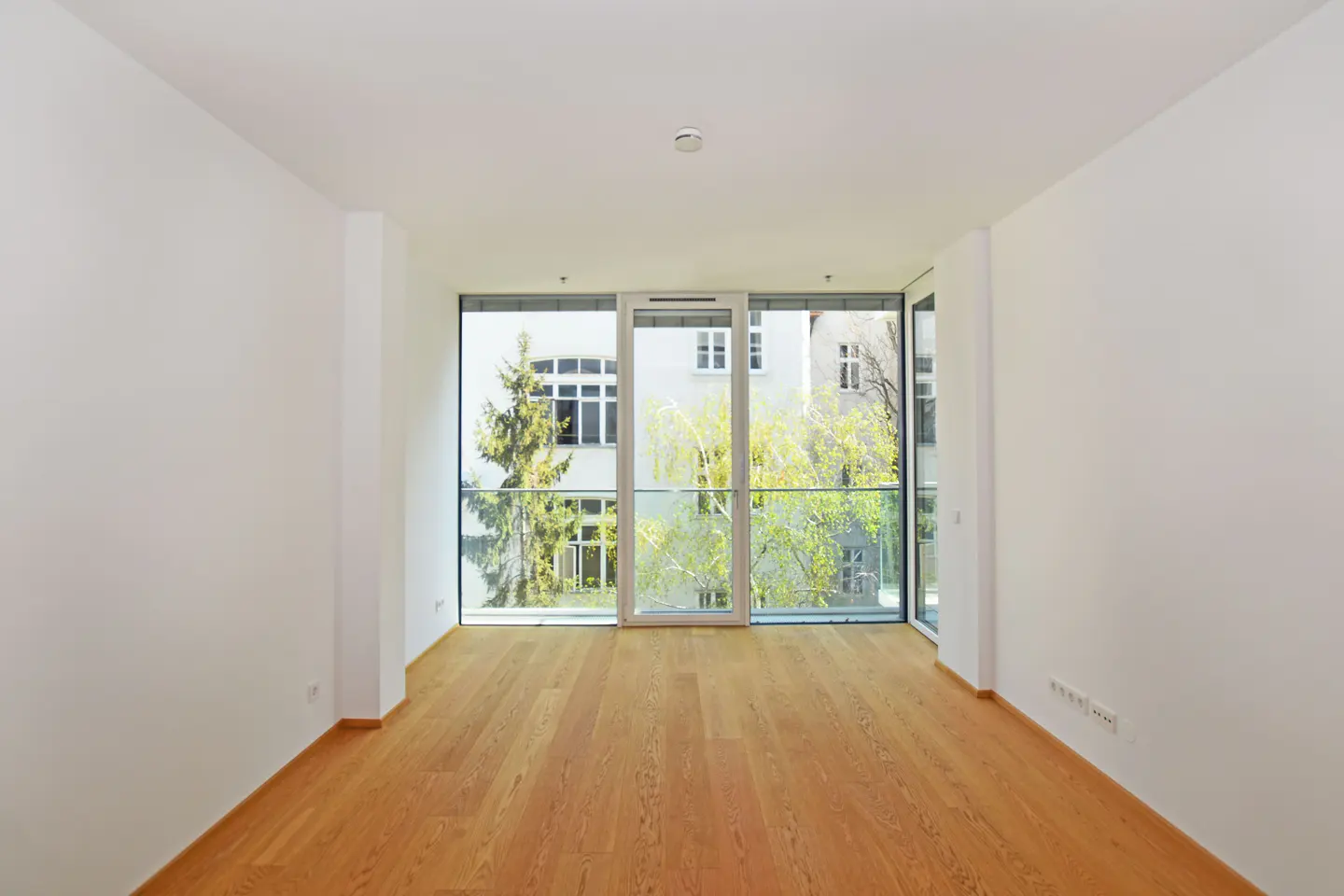 Empty room with light wood floors and white walls. Large windows overlook trees and a building.