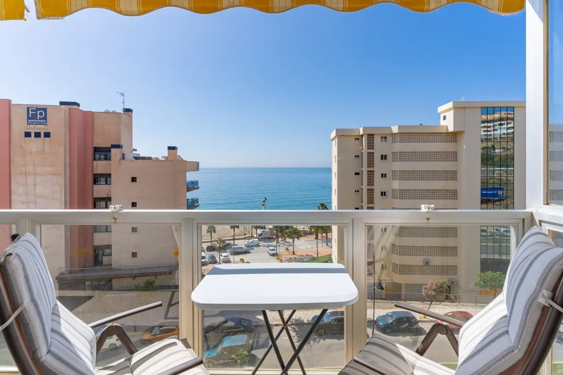 Balcony view with a small white table and two striped chairs overlooking a coastal street, beach, and the blue sea.
