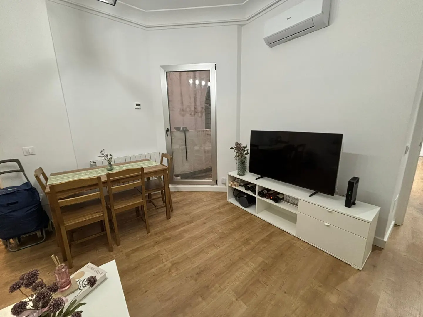 Modern living room with wood floors, a dining set, a white TV stand with a flat-screen TV, and a glass balcony door.