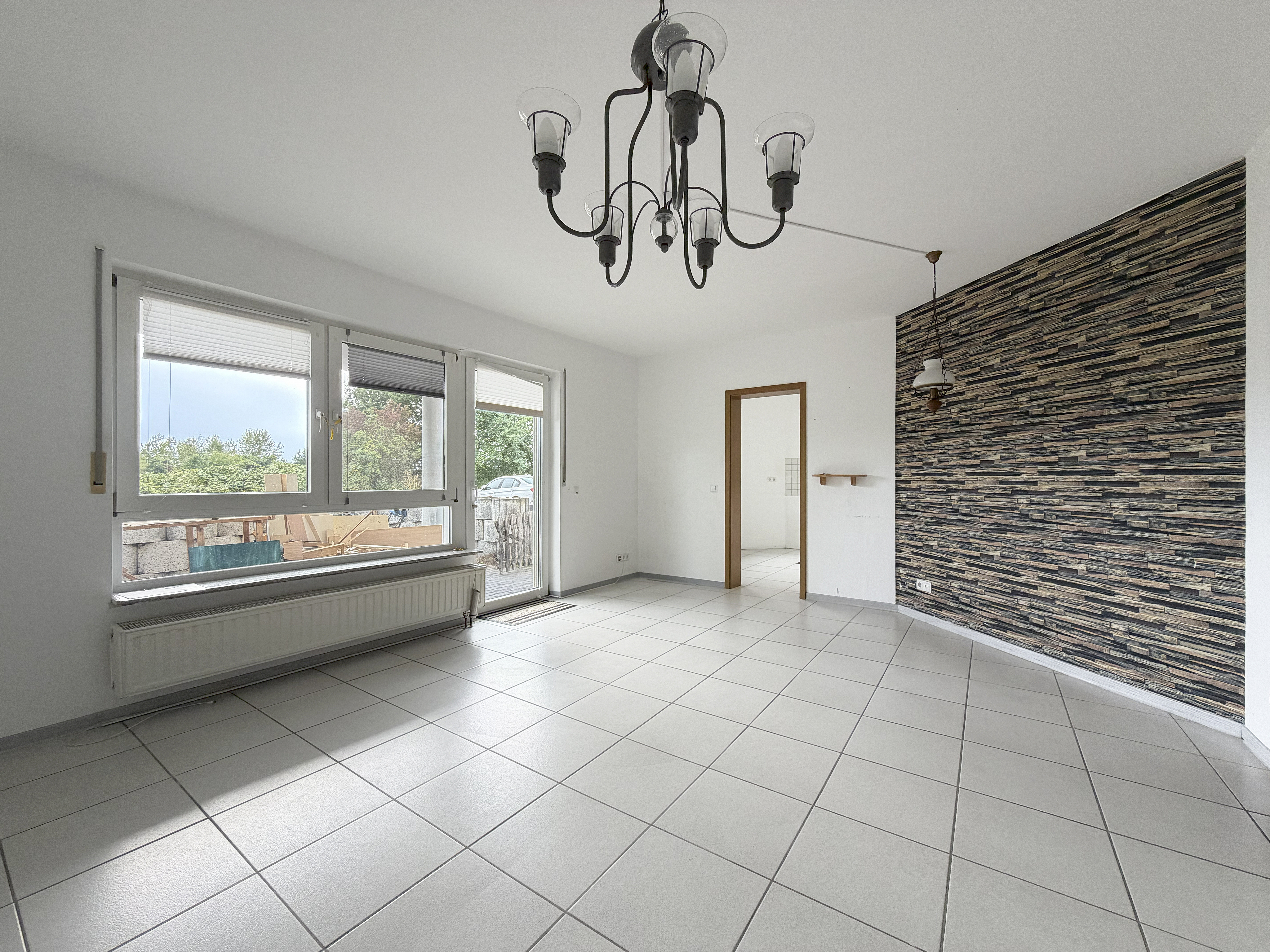 Bright room with white tiled floors, a black chandelier, and a textured stone-look accent wall, featuring large windows. [AI-generated text]