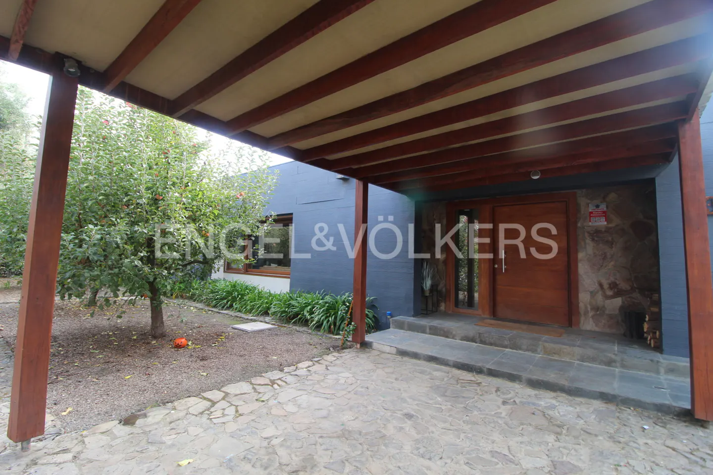 Exterior view of a modern house with a stone driveway and wooden carport. The house has a gray facade and a wooden front door.
