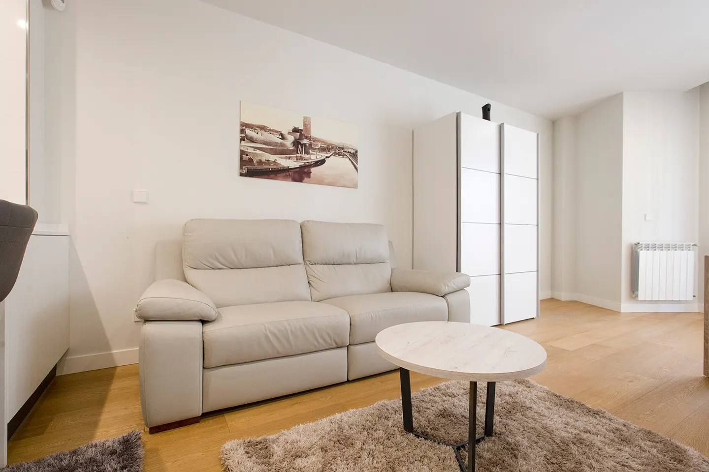 Living room with a beige leather sofa, round table on a shag rug, and a white wardrobe against a white wall.