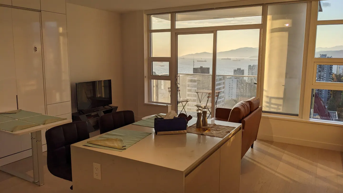 Condo interior with a white kitchen island, brown leather couch, and a large window overlooking a city and ocean view.