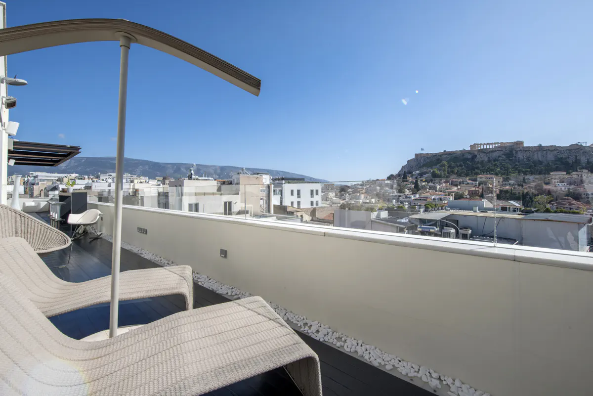 Rooftop patio with lounge chairs and a view of Athens, Greece. The Acropolis is visible in the distance under a clear blue sky.
