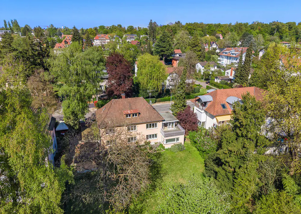 Aerial view of a two-story beige house with a red tile roof, surrounded by lush green trees and other houses.