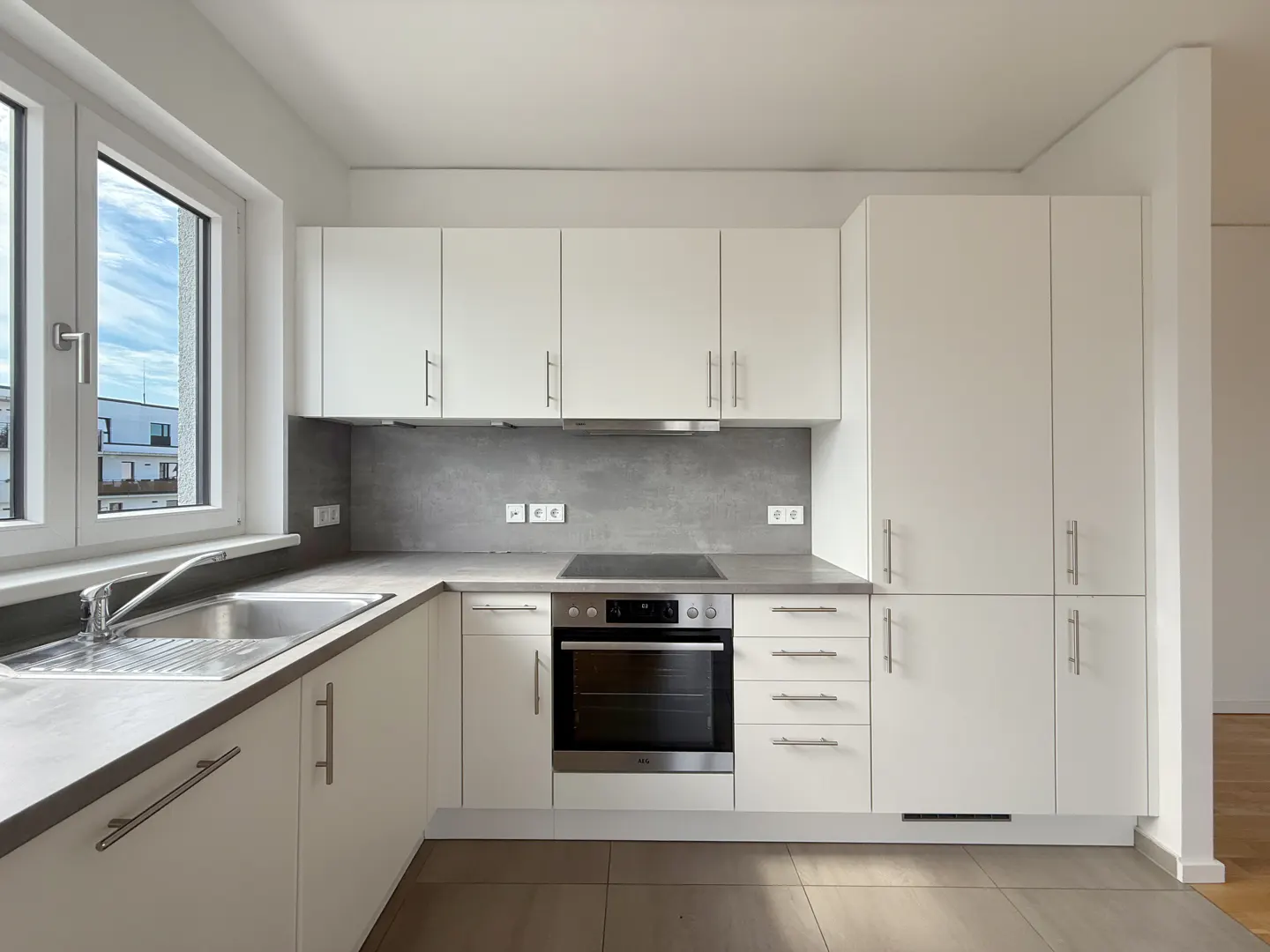 Bright kitchen with white cabinets, gray countertops, and stainless steel appliances. A window overlooks a building and blue sky.