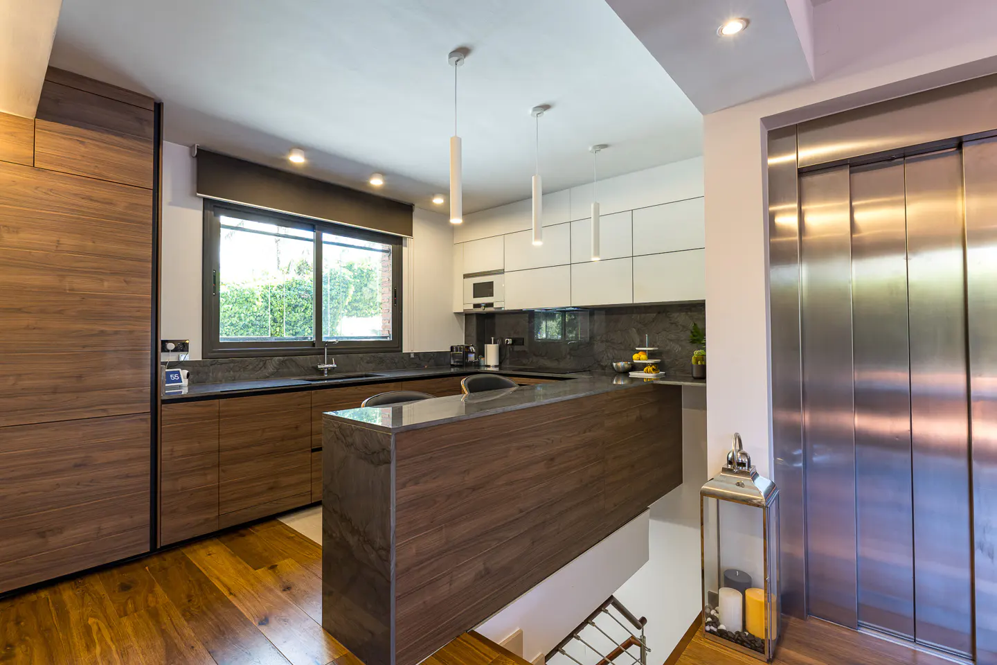 Modern kitchen with wood floors, cabinets, and island. Stainless steel elevator is visible on the right.
