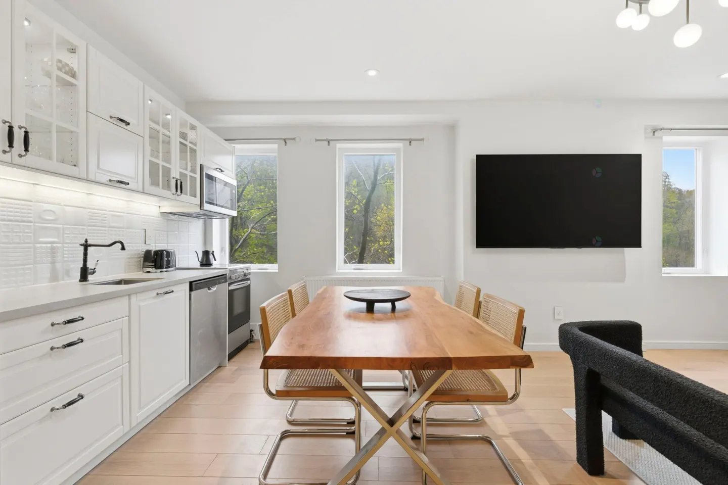 Bright, modern kitchen and dining area with white cabinets, wood table, and stainless steel appliances. Windows overlook trees.