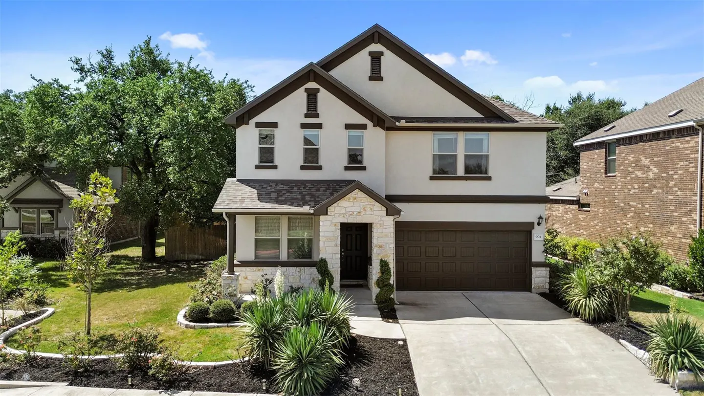 Two-story house with white stucco, brown trim, and stone accents. Landscaped front yard with a concrete driveway. Blue sky background.