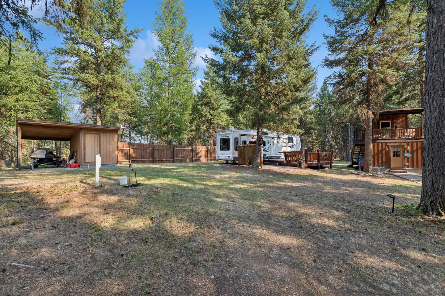 A wooded property with a brown cabin, RV, shed, and a wooden fence under a blue sky.