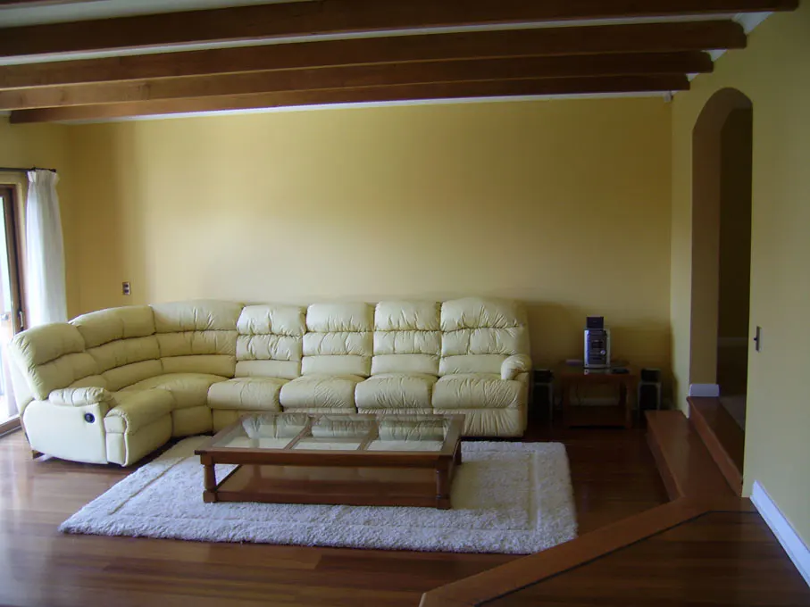 Living room with yellow walls, wood floors, and exposed beams. A cream-colored sectional sofa sits on a white rug in front of a glass-topped coffee table.