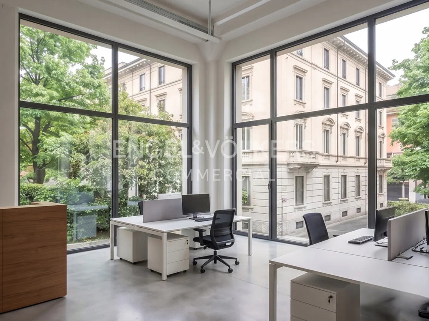 Bright office space with large windows overlooking greenery and a building. White desks and black chairs furnish the room.