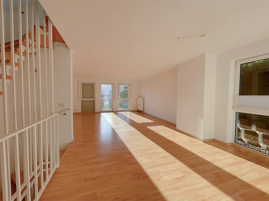 Bright, empty living room with wood floors, white walls, and a staircase with white railings. Sunlight streams through windows and doors.