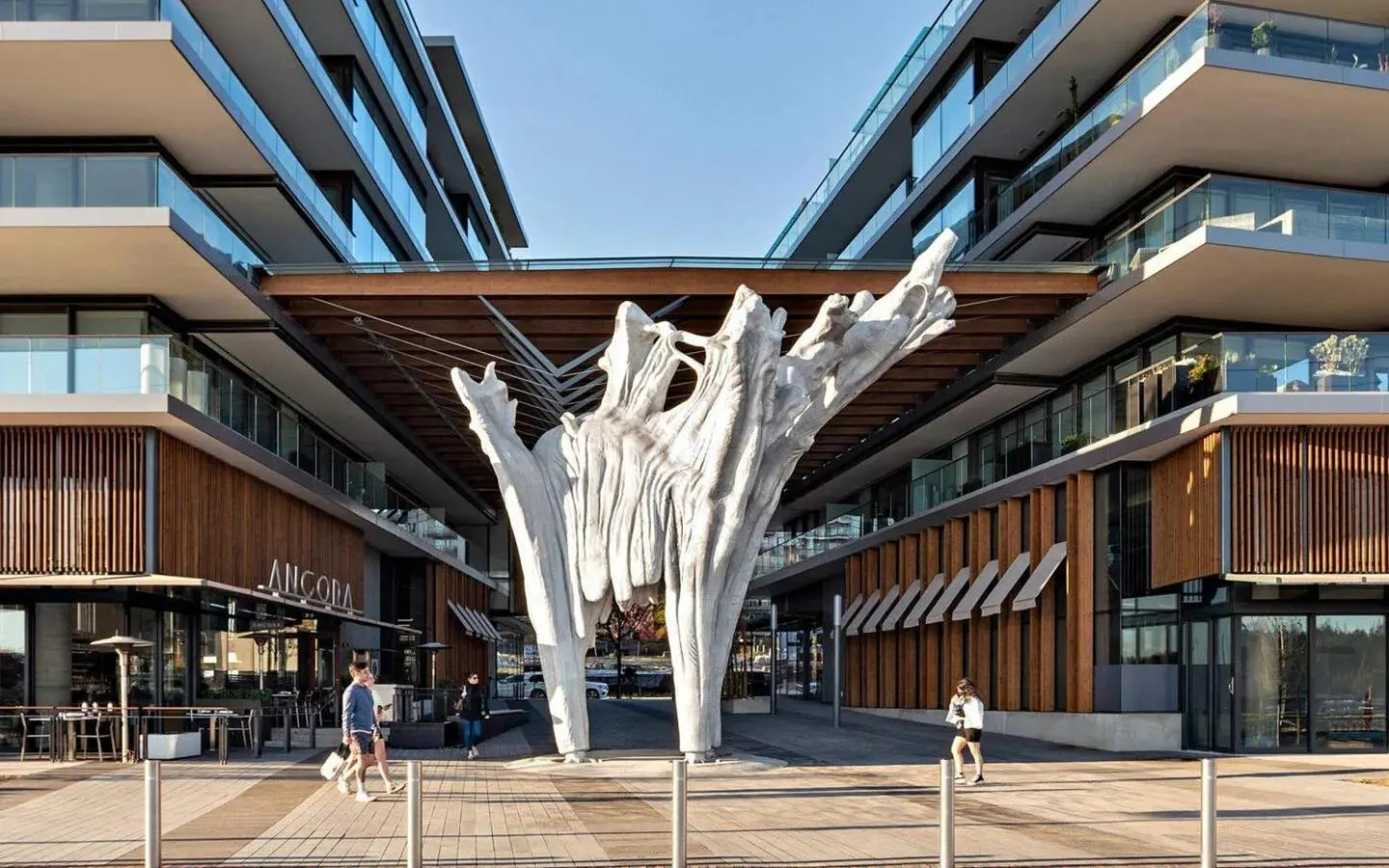 Modern apartment complex with a large white tree sculpture in the center courtyard. People walk along the brick-paved path.