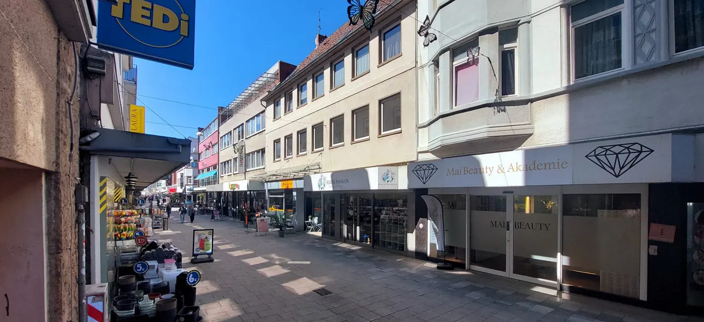 Street view of shops and buildings on a sunny day. "Mai Beauty & Akademie" is visible.